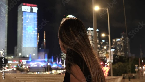 Night shot of huge Israel flag hanging on Azrieli tower with a young concerned jewish woman looking at it from a bridge. Big city lights shot of a worried lady with a focus on Israel flag background. photo