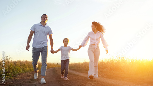 Parents lift daughter taking hands walking in field at sunset. Sun rays fall on happy family, sunlight