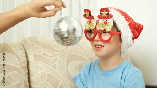 preparing for Christmas. A boy in a red Santa hat is playing with Christmas balls sitting on the couch.