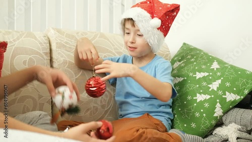 A cheerful smiling boy in a red Santa hat is playing with Christmas balls sitting on the sofa.