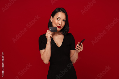 Thoughtful asian woman using mobile phone and credit card isolated over red background