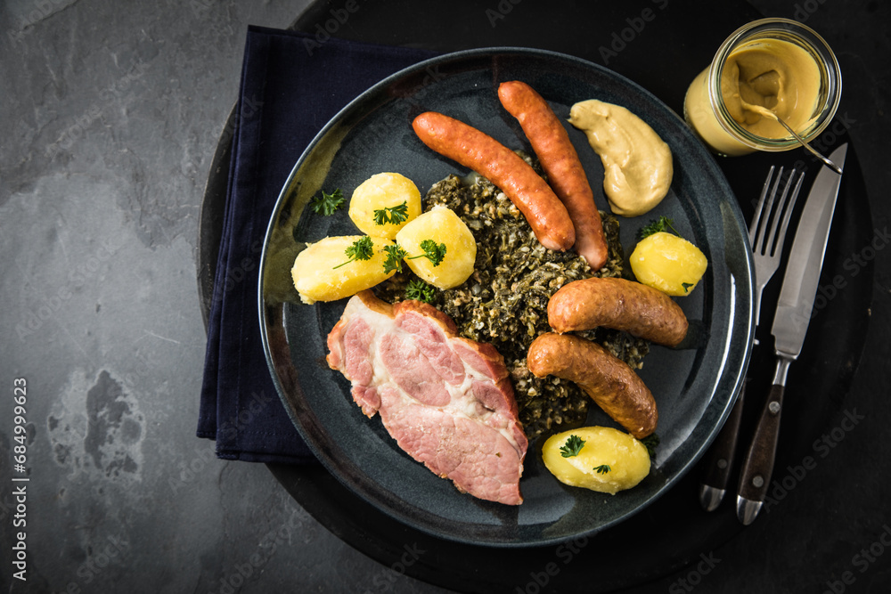 Oldenburger Grünkohl mit geräucherter Kohlwurst, Pinkel, Kassler Nacken, Kartoffeln, Senf, Petersilie auf Teller und dunkel schwarz Schiefer Hintergrund Oldenburger Grünkohl mit geräucherter Kohlwurst, Pinkel, Kassler Nacken, Kartoffeln, Senf, Petersilie auf Teller und dunkel schwarz Schiefer Hintergrund