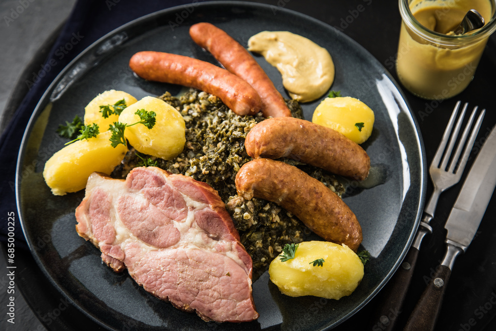 Oldenburger Grünkohl mit geräucherter Kohlwurst, Pinkel, Kassler Nacken, Kartoffeln, Senf, Petersilie auf Teller und dunkel schwarz Schiefer Hintergrund Oldenburger Grünkohl mit geräucherter Kohlwurst, Pinkel, Kassler Nacken, Kartoffeln, Senf, Petersilie auf Teller und dunkel schwarz Schiefer Hintergrund