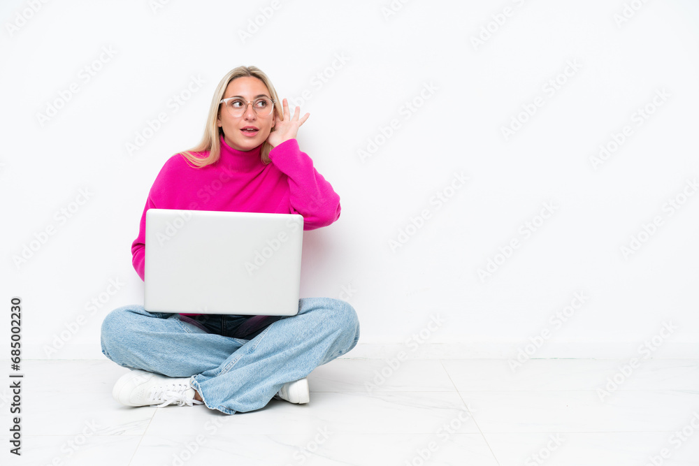 Fototapeta premium Young caucasian woman with laptop sitting on the floor listening to something by putting hand on the ear