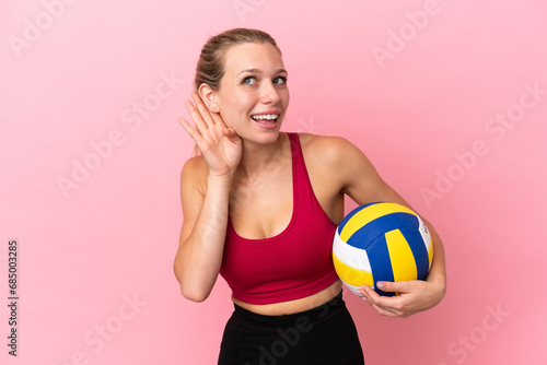 Young caucasian woman playing volleyball isolated on pink background listening to something by putting hand on the ear