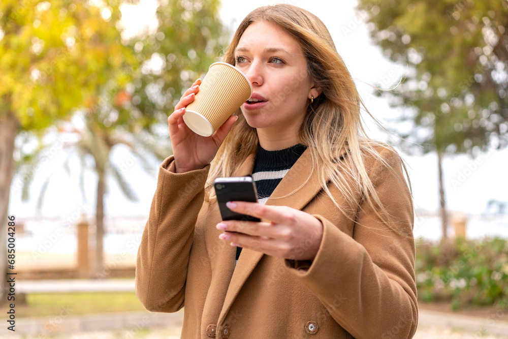 Young pretty blonde woman using mobile phone and holding a take away coffee at outdoors