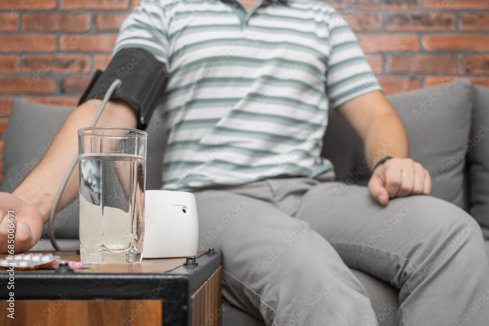 Defocused background with man sitting on sofa at home and measures blood pressure on hand with modern digital tonometer, focus on blister pack with pills on foreground Defocused background with man sitting on sofa at home and measures blood pressure on hand with modern digital tonometer, focus on blister pack with pills on foreground