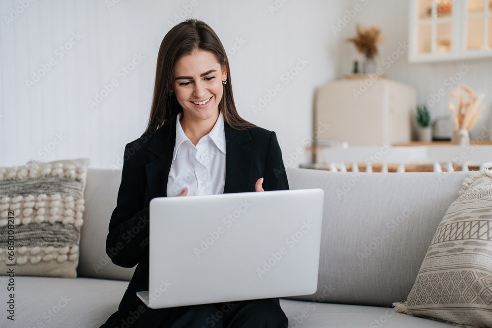 Elegant young businesswoman in suit talks via internet using laptop at home sitting on couch toothy smiles. Professional lawyer remote consulting client. Remote work, home office. Elegant young businesswoman in suit talks via internet using laptop at home sitting on couch toothy smiles. Professional lawyer remote consulting client. Remote work, home office.