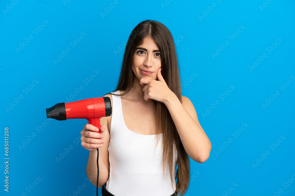 Young caucasian woman holding a hairdryer isolated on blue background thinking Young caucasian woman holding a hairdryer isolated on blue background thinking