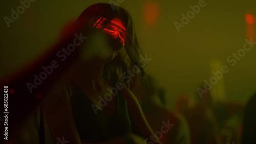 Cheerful girl dancing and jumping on dance floor enjoying party in nightclub at music festival. Young woman in glowing glasses hangs out in club with multi-color neon illumination, crowd in background