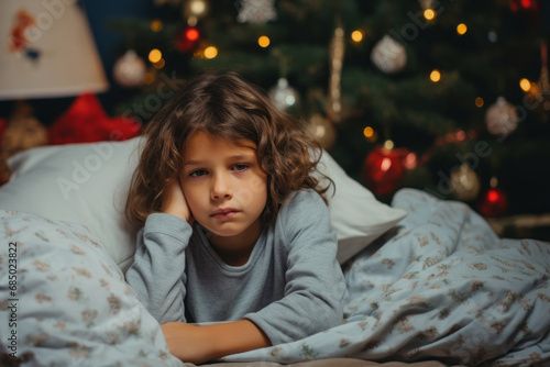 Sad Christmas at home. Sick child lies in bed next to a decorated Christmas tree at home. Kid is waiting for Santa Claus presents © vejaa
