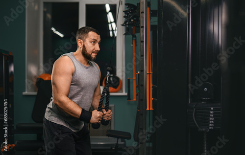 A swarthy man with a beard with effort performs a triceps exercise pulling the block to the bottom in the gym.