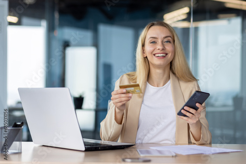 Portrait of a young businesswoman sitting in the office at a desk  smiling at the camera  holding a credit card and a phone