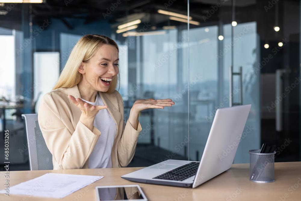 Fototapeta premium A happy young businesswoman works in the office, sits at the table and smilingly talks at an online meeting with clients and partners via video call, rejoices at success, a concluded agreement