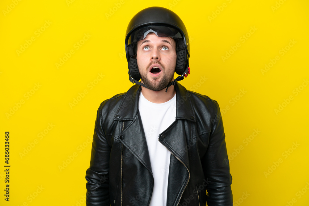Young caucasian man with a motorcycle helmet isolated on yellow background looking up and with surprised expression Young caucasian man with a motorcycle helmet isolated on yellow background looking up and with surprised expression
