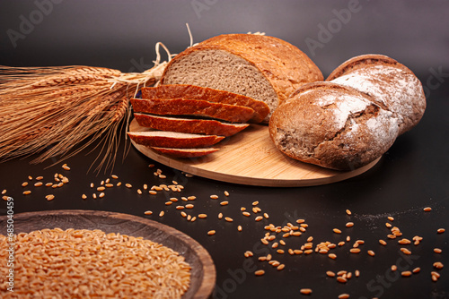 Whole and sliced breads, wheat ears and scattered wheat grains on wooden plate on black background. Photograph. (ID: 685040459)