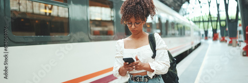 Close-up portrait of a young woman in glasses with a smartphone and backpack stands at the station. Positive woman using mobile phone outdoors in urban background.