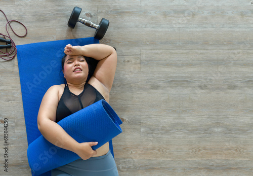 A plus-size woman lies exhausted on a blue mat, taking a breather after a vigorous workout session with dumbbell and jump rope. Top view photo