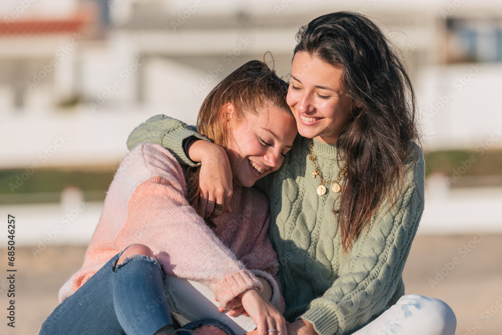Happy girlfriends sitting and embracing on street against blurred buildings in daylight Happy girlfriends sitting and embracing on street against blurred buildings in daylight