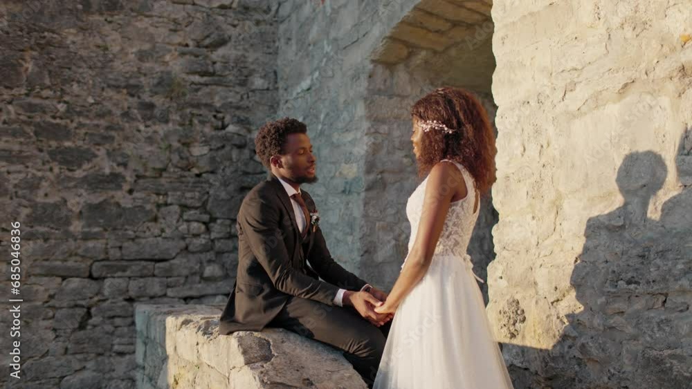 African American groom sitting on stone while holding hand of his bride before castle. Young couple talking promises of love, fidelity, and words of support to each other. Wedding vows. Happy together African American groom sitting on stone while holding hand of his bride before castle. Young couple talking promises of love, fidelity, and words of support to each other. Wedding vows. Happy together
