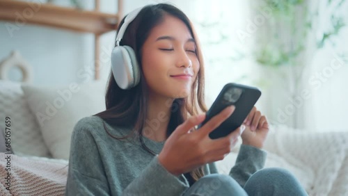 Happy asian woman listening to music from mobile phone while sitting on the rug beside to the sofa at homes, Smiling girl relaxing with headphones in morning, Time to relax. photo