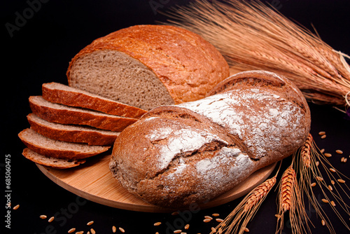 Whole and sliced breads, wheat ears and scattered wheat grains on wooden plate on black background. Photograph. (ID: 685060475)