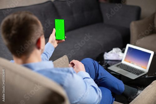 Male psychologist sitting at workplace and looking at mobile phone screen with green chromakey. Operating laptop with image on desktop