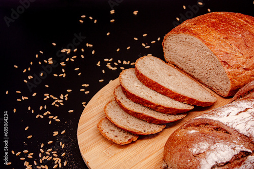 Whole and sliced breads, wheat ears and scattered wheat grains on wooden plate on black background. Photograph. (ID: 685064234)