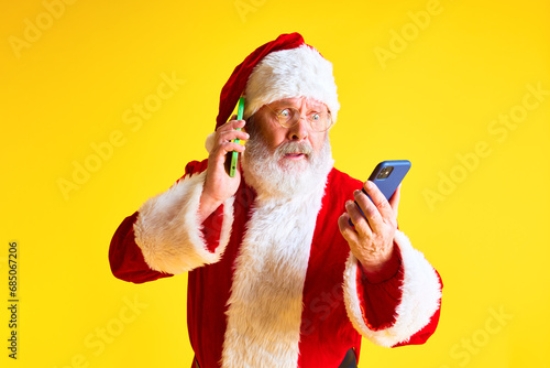 Portrait of senior confused man with long beard receives calls from two phones from children who are waiting for gifts against yellow studio background. photo