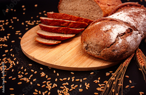 Whole and sliced breads, wheat ears and scattered wheat grains on wooden plate on black background. Photograph. (ID: 685068419)