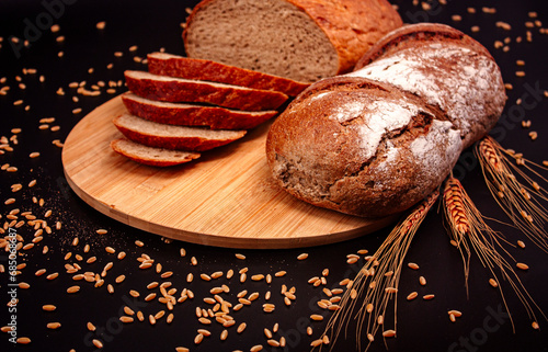 Whole and sliced breads, wheat ears and scattered wheat grains on wooden plate on black background. Photograph. (ID: 685068687)