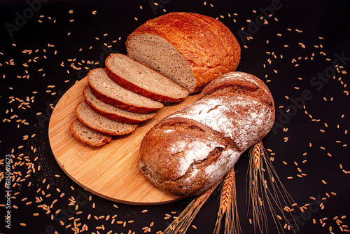 Whole and sliced breads, wheat ears and scattered wheat grains on wooden plate on black background. Photograph. (ID: 685069623)