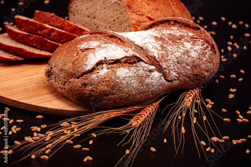 Whole and sliced breads, wheat ears and scattered wheat grains on wooden plate on black background. Photograph. (ID: 685073006)