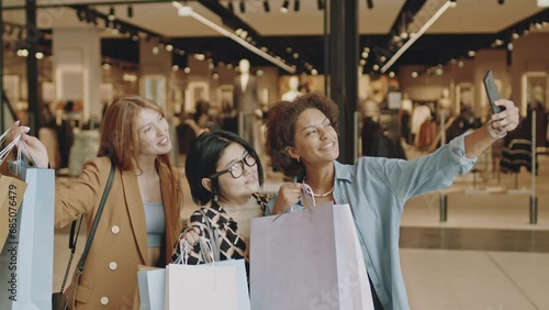 Medium shot of three happy multiethnic female friends taking selfie with paper shopping bags standing against modern clothing store in big department store photo