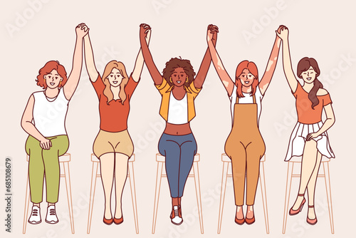 Team of diverse women raise hands together as sign of unity and cohesion among female community. Girls of different races dressed in casual clothes sit on chairs and demonstrate high team spirit
