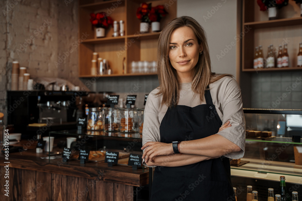 Smiling woman in apron standing confidently in a cozy cafe environment, with shelves of flowers in the background Smiling woman in apron standing confidently in a cozy cafe environment, with shelves of flowers in the background