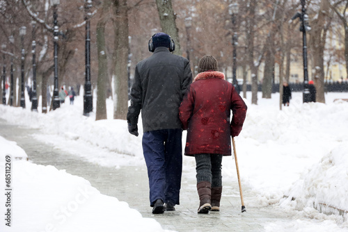 Elderly woman witj cane and man walking in winter park, rear view. Old couple in warm clothes during snow weather, concept of old age