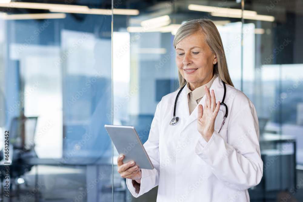 An older gray-haired woman is standing in the office of the clinic in a coat, holding a tablet in her hands. chats on a video call, conducts an online consultation, greets and waves at the camera. An older gray-haired woman is standing in the office of the clinic in a coat, holding a tablet in her hands. chats on a video call, conducts an online consultation, greets and waves at the camera.