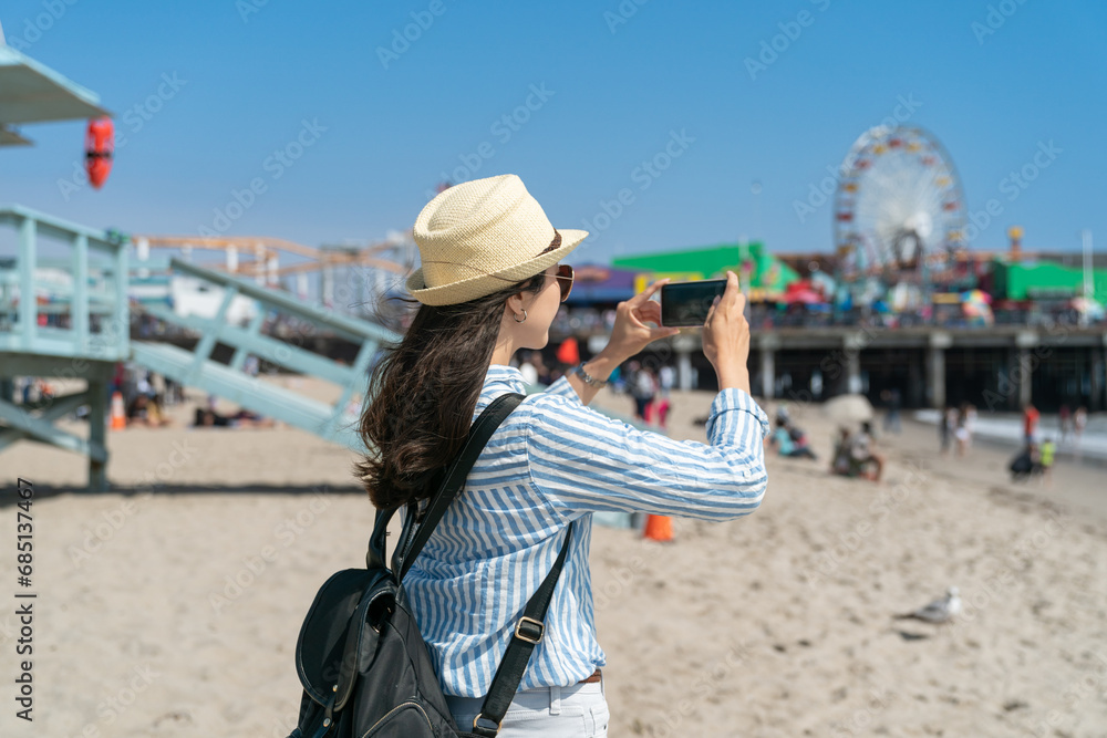 Fototapeta premium rear view of asian Japanese girl backpacker taking photos with smartphone on beach of santa monica pier near Lifeguard Tower