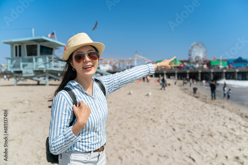 happy asian Taiwanese lady visitor looking at camera and pointing finger at ferris wheel in distance near Lifeguard Tower at santa monica beach