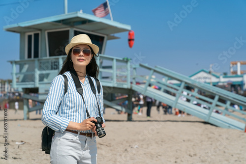 fashionable asian Korean girl photographer on vacation looking at beautiful scenery at santa monica beach with lifeguard tower at background