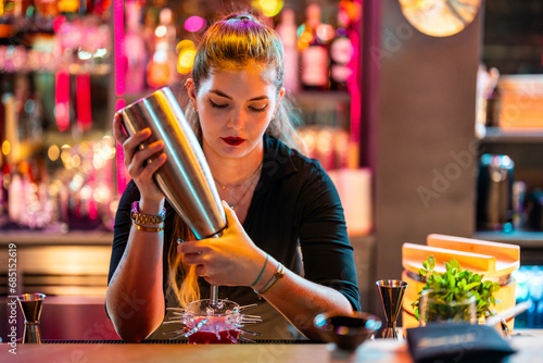Focused young female barmaid with blonde hair standing at counter while making cocktail at illuminated bar