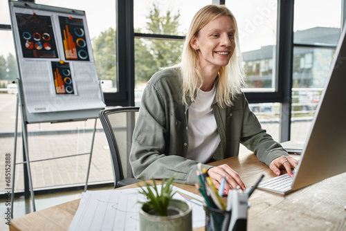 joyous non binary person working at table in office and smiling cheerfully, business concept photo