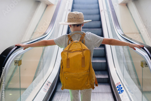 Back view of anonymous boy wearing backpack and straw hat with arms outstretched standing on moving escalator at airport photo