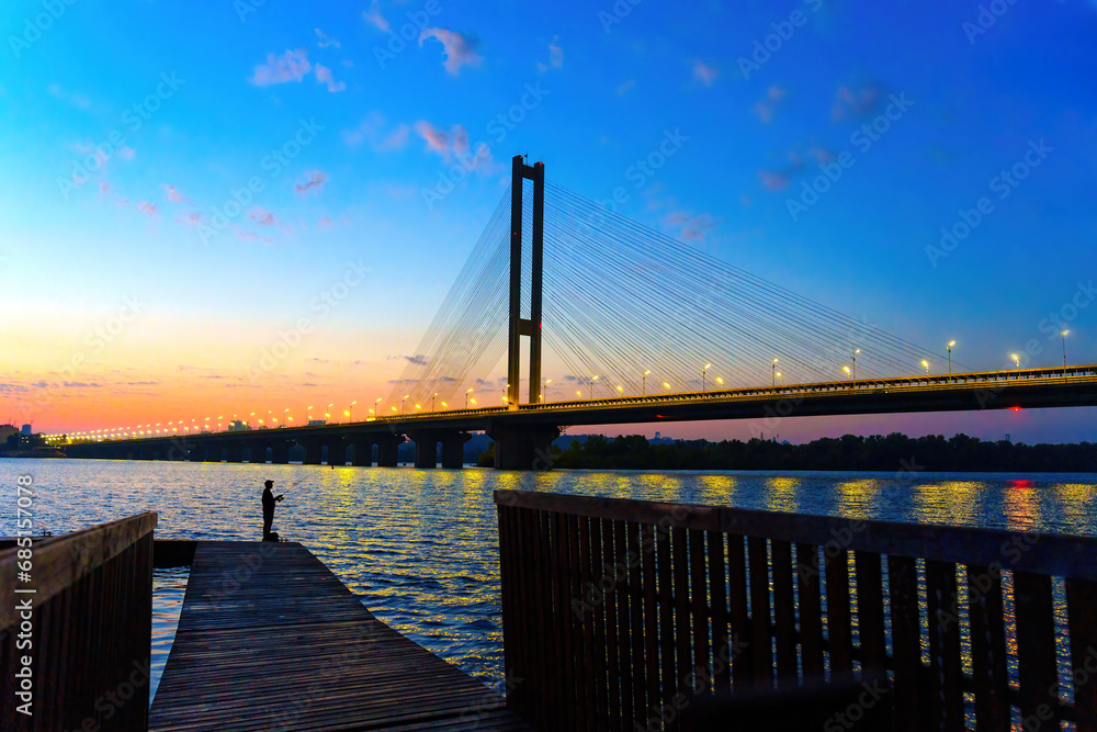 Naklejka premium Silhouetted Angler on a Wooden Pier against a Cable-Stayed Bridge