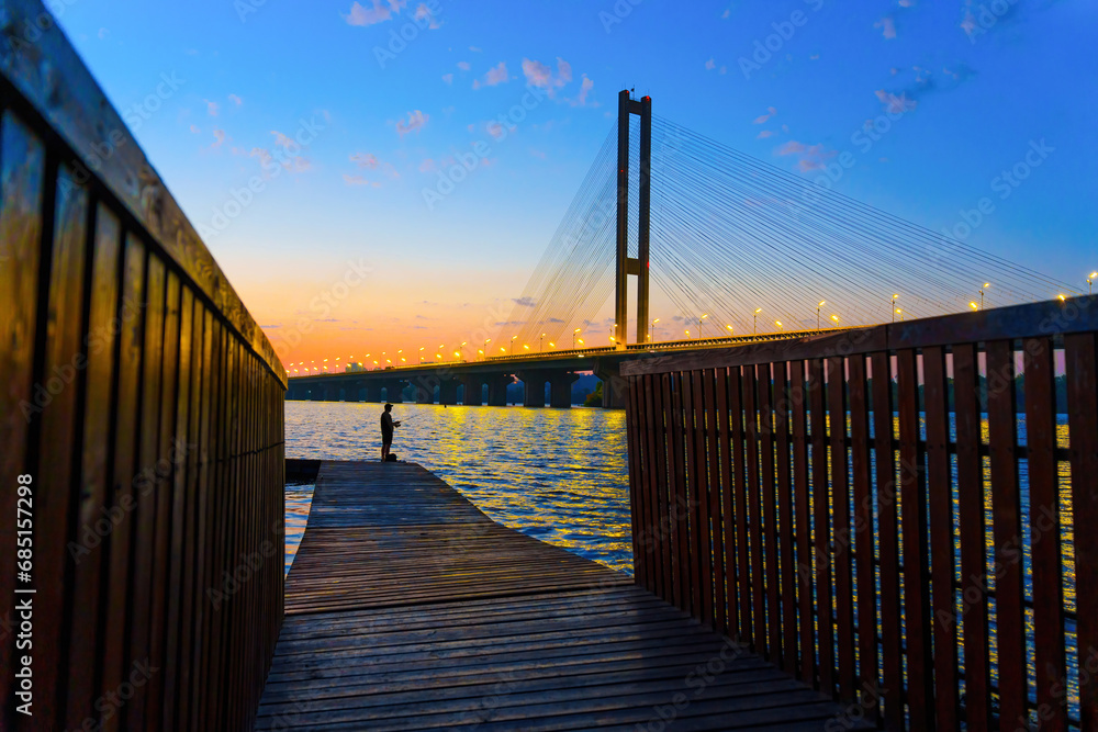 Naklejka premium Wooden Dock and Fisher with a Cable-Stayed Bridge at Sunset