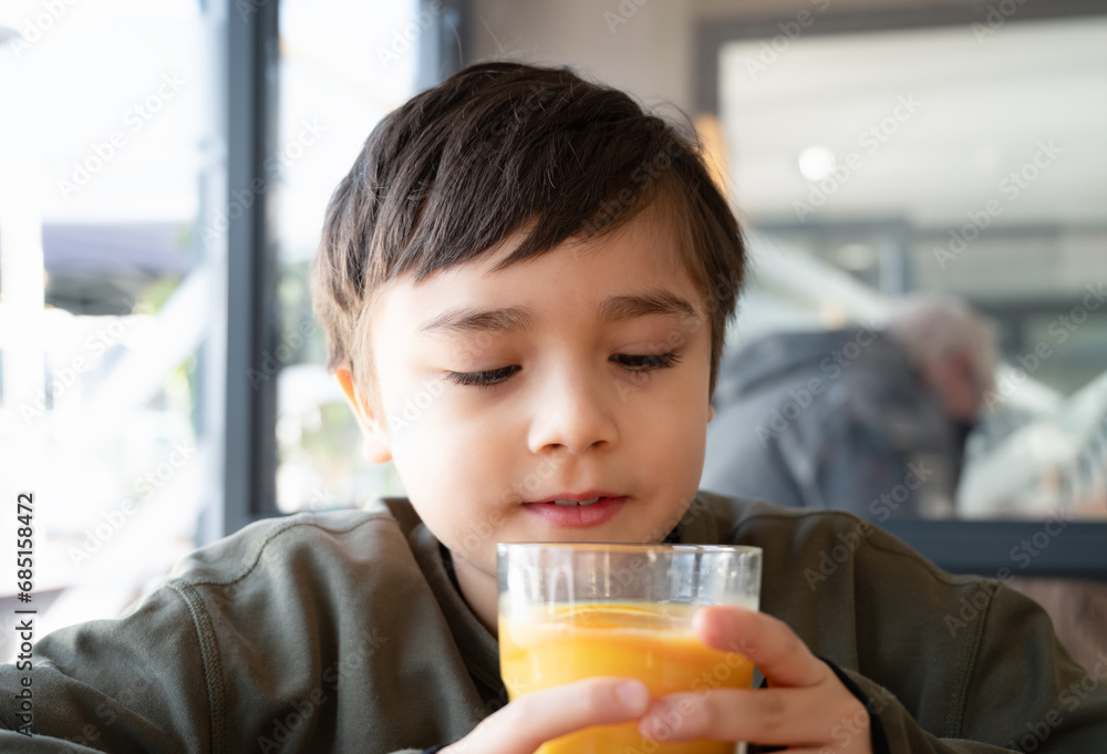 Portrait Young kid boy drinking fresh orange juice for breakfast in cafe, Happy child holding glass of fruit juice while waiting for food in restaurant, Healthy food lifestyle concept Portrait Young kid boy drinking fresh orange juice for breakfast in cafe, Happy child holding glass of fruit juice while waiting for food in restaurant, Healthy food lifestyle concept
