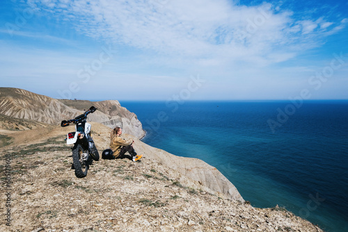 Female rider relaxing in enduro offroad motorcycle travel on mountain top, beautiful sea shore and mountains landscape on background © Annatamila