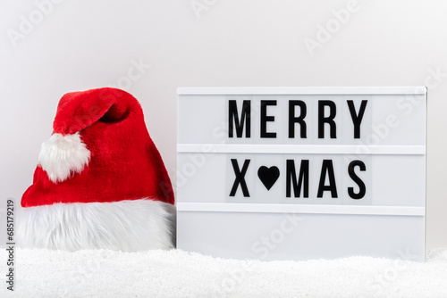 White lightbox with text MERRY XMAS and Santa Claus hat on a table with snow. Minimal Christmas or New Year concept.