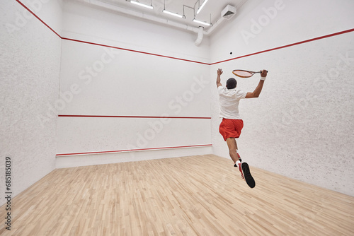 athletic african american player with racquet jumping and playing squash in court, back view